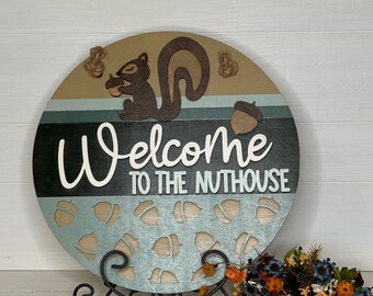 May include: Round wooden sign with a squirrel holding an acorn, the text "Welcome to the Nuthouse", and acorn cutouts. The sign has a brown, green, and blue color scheme, with rope for hanging.
