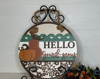 May include: Round wooden sign with a fall theme. The sign features a pumpkin design and the words "HELLO Gourd-geous". It has a decorative border and is held by a black metal stand with a rope hanger.