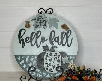 May include: A round wooden sign with the words "hello fall" in black script. The sign features a pumpkin design and decorative leaves, with twine accents. The sign is displayed on a black metal stand, with a bouquet of flowers in the foreground.