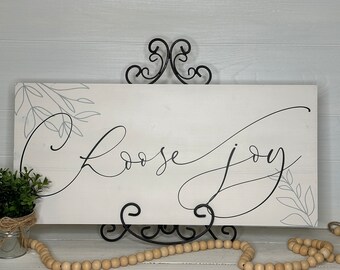 May include: A rectangular wooden sign with the words "Choose Joy" in elegant black script. The sign is white with gray leaf accents and is displayed on a black metal stand. A small potted plant and wooden beads are in the foreground.