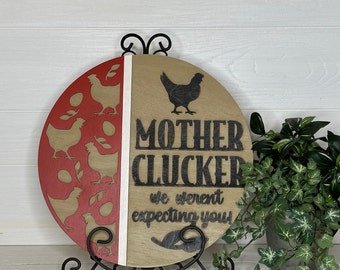 May include: A round wooden sign with a red and tan color scheme. The left side features laser-cut chicken and egg designs, while the right side displays the words "MOTHER CLUCKER we weren't expecting you." The sign is displayed on a black metal stand.