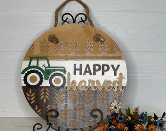 May include: A round wooden sign with a rope handle, featuring a tractor and the words "HAPPY harvest." The sign has a wheat design and is painted in shades of brown, white, green, and gray. It is displayed on a black metal stand.