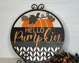 May include: A round wooden sign with a pumpkin design and the words "HELLO Pumpkin". The sign features an orange pumpkin with a green stem and leaf, set against a black and white background. The sign is displayed on a black metal stand.