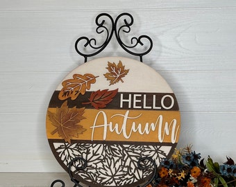 May include: A round wooden sign with the words "HELLO AUTUMN" in white lettering. The sign features a design of fall leaves in shades of brown, orange, and cream. It is displayed on a black metal stand, with a white wooden background.