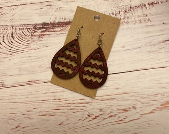 May include: A pair of teardrop-shaped wooden earrings with a zigzag pattern cut out. The earrings are a deep red color with silver-colored hooks. They are displayed on a brown card with a silver-colored fastener, set against a white and light brown wooden surface.