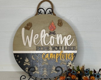 May include: Round wooden sign with the word "Welcome" and the phrase "Hope you like campfires." The sign features a campfire design and is decorated with rope and a black metal hanger. The sign is displayed on a black metal stand.
