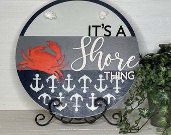 May include: A round wooden sign with a nautical theme. The sign features a red crab, white anchors, and the phrase "IT'S A Shore THING". The sign is supported by a black metal stand and has two small white rope accents.