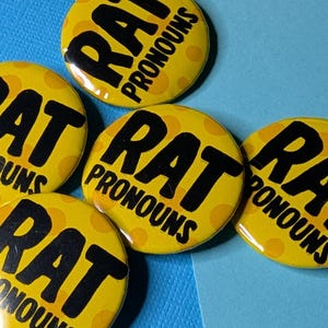 May include: Five yellow round buttons with the text "RAT PRONOUNS" in black. The buttons have a polka dot pattern and are arranged on a blue surface.