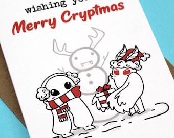 May include: A white greeting card with the text "wishing you a Merry Cryptmas" in red and black. The card features cartoon illustrations of a snowman, a creature with a gift, and a creature wearing a scarf and earmuffs. The card is inside a brown envelope.