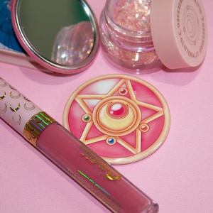 May include: A pink and gold makeup compact with a star and crescent moon design. The compact is surrounded by a pink and white makeup brush, a pink and white compact mirror, and a jar of pink glitter.