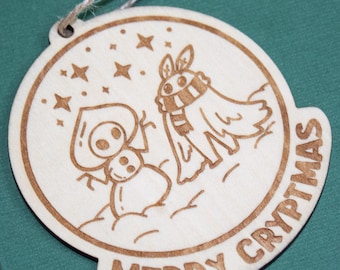May include: Wooden Christmas ornament with a whimsical design. The ornament features a snowman and a rabbit wearing a scarf, with stars in the background. The words "MERRY CRYPTMAS" are etched around the edge. A jute string is attached for hanging.