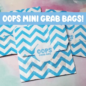 May include: Three blue and white chevron patterned paper bags with a white circle sticker that says "Oops Grab Bag" on one of the bags. The bags are labeled "Oops Mini Grab Bags!"