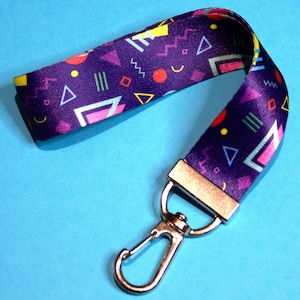 May include: A purple lanyard with a geometric pattern of triangles, zigzags, and circles in yellow, pink, green, and orange. The lanyard has a silver-colored metal clasp and a rectangular metal piece.