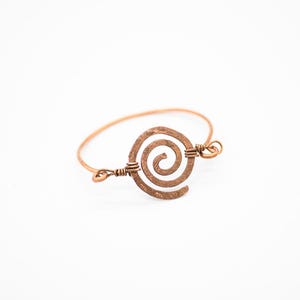 May include: Copper-toned bracelet featuring a spiral design at the center. The bracelet has a simple band with a decorative spiral element, showcasing a handcrafted aesthetic. The metal has a warm, reddish-brown hue.