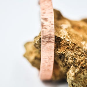 May include: A copper-colored, textured bracelet is displayed against a rough, tan-colored rock. The bracelet has a rectangular shape with a detailed, etched surface. The background is a soft white.