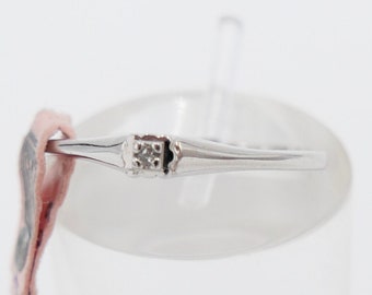 Könnte beinhalten: Ein silberner Ring mit einem kleinen Diamanten in der Mitte. Der Ring wird auf einem weißen Ständer mit einem rosa Etikett präsentiert, auf dem die Nummer 1046 steht.