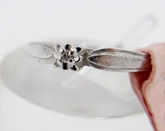 Könnte beinhalten: Ein silberner Ring mit einem kleinen Diamanten, der in einem blumenförmigen Design gefasst ist. Der Ring liegt auf einer weißen Oberfläche mit einem rosa Etikett, auf dem "18/7 B51 M2" steht.