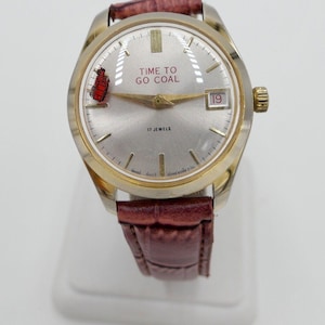 May include: A silver and gold wristwatch with a brown leather strap. The watch face has a white background with black markings and a red and white image of a train engine. The text "TIME TO GO COAL" is printed in red on the watch face. The date window shows the number 19.