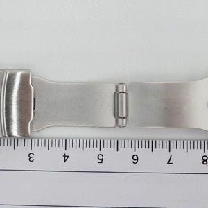 May include: A silver watch band with a clasp and a measurement ruler in the background.
