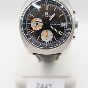 May include: A vintage Tissot Automatic Navigator watch with a black dial, silver case, and black leather strap. The watch features chronograph subdials, orange accents, and a date display at the 3 o'clock position. The watch is displayed on a white stand.