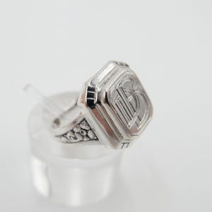 May include: A silver signet ring with an ornate design and a square face featuring the letter "B".