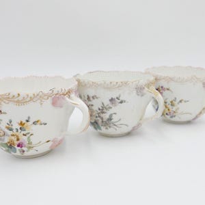 May include: Three white porcelain teacups with floral designs and gold trim. The cups have a scalloped edge and a delicate floral pattern in shades of pink, yellow, and green.