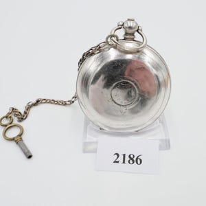 May include: A silver pocket watch with a chain and key. The watch has an engraved design on the back and the number 2186 is visible on a white tag.