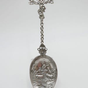 May include: A silver spoon with a detailed design of a windmill and a landscape on the bowl. The handle is ornate and features a crown and two lions.