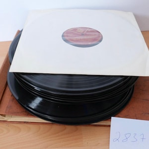 Könnte beinhalten: Ein Stapel schwarzer Schallplatten liegt auf einem braunen, strukturierten Album-Aufbewahrungsordner. Die oberste Platte ist teilweise von einer weißen Papierhülle mit einem kreisförmigen Ausschnitt bedeckt, der ein rot-weißes Etikett freigibt.