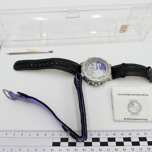 Può includere: Un orologio da polso argento e nero con quadrante bianco e cinturino viola. L'orologio ha un display digitale e un cinturino in pelle nera. L'orologio è in una custodia di plastica trasparente con un piccolo cacciavite.