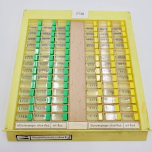 May include: A yellow plastic tray with a wooden divider contains small clear plastic containers with watch hands. The containers are labeled with measurements in millimeters. The tray is labeled "1730" and has the text "Minutenzeiger ohne Rad. mit Rad." and "Stundenzeiger ohne Rad. mit Rad."  The tray also has the text "Dauphinform mit+ohne R."