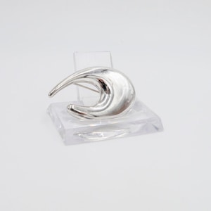 Puede incluir: Un broche de plata en forma de media luna abstracta. El broche está hecho de metal liso y curvo y tiene un diseño simple y moderno.
