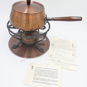 May include: Copper fondue pot with a wooden handle and a black metal stand. The pot has a lid and a decorative pattern. The stand has a copper base and a small burner. The image also includes a sheet of paper with the text "4 Rezepte" and a recipe for "Rinds Fondue".