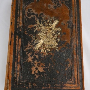 Può includere: Libro antico con una copertina in pelle marrone usurata e bordi neri. La copertina presenta un disegno dorato in rilievo e un bordo decorativo. Il dorso del libro è visibile e i bordi delle pagine sono invecchiati. Il numero "2586" è su un'etichetta bianca.