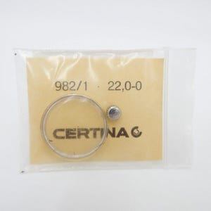 May include: A close-up of a small plastic bag containing a clear watch crystal and a silver watch crown. The bag is labelled with the text "982/1 22,0-0 CERTINA C".