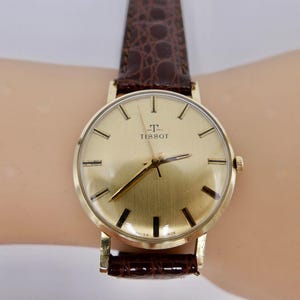 Può includere: Un orologio da polso dorato con un cinturino in pelle marrone. Il quadrante dell'orologio è rotondo e ha una lunetta dorata. Il quadrante dell'orologio ha indici orari romani neri e un logo TISSOT dorato.