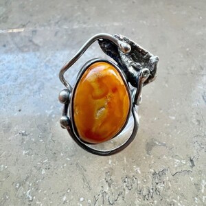 Könnte beinhalten: Ein silberner Ring mit einem Bernstein, der in einem einzigartigen, abstrakten Design gefasst ist. Der Ring hat ein strukturiertes, organisch geformtes Band mit einem großen, ovalen Bernstein.