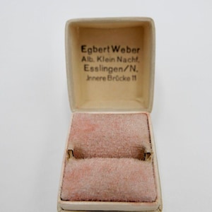 Puede incluir: Una caja de anillo blanca con un interior de terciopelo rosa. La caja tiene el texto "Egbert Weber Alb. Klein Nachf. Esslingen/N. Innere Brücke 11" impreso en el exterior.