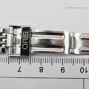 以下が含まれることがあります： メッシュデザインのシルバーカラーの時計バンド。バンドには「ORIS」のブランド名が刻印された留め金が付いています。定規が下に置かれ、センチメートル単位で測定されています。