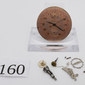 May include: A disassembled watch face with a copper-coloured dial and black hands. The watch face has a tachymeter scale and a small sub-dial. The watch face is surrounded by a clear plastic stand. The watch parts are scattered on a white surface. The number 160 is written on a white piece of paper.