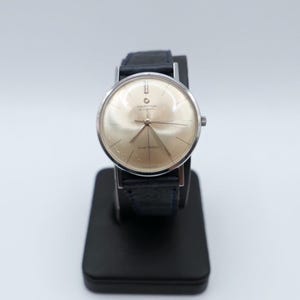 Könnte beinhalten: Eine Vintage Certina Blue Ribbon Automatik-Armbanduhr mit goldfarbenem Zifferblatt und silberfarbenem Gehäuse. Die Uhr hat ein schwarzes Lederarmband und wird auf einem schwarzen Ständer präsentiert. Das Zifferblatt trägt die Aufschrift "Certina" und "Blue Ribbon".