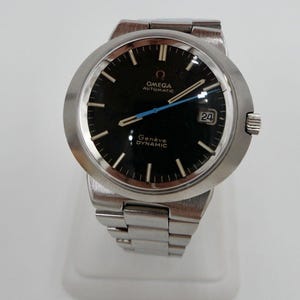 May include: A silver Omega Geneve Dynamic automatic wristwatch with a black face and a silver metal band. The watch face has silver hour markers and hands, a blue second hand, and a date window at the 3 o'clock position.