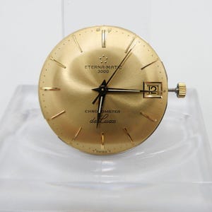 Puede incluir: Esfera de reloj dorada con agujas y marcas negras. La esfera dice "Eterna-Matic 3000 Chronometer de Luxe" y "Swiss T".