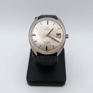 Könnte beinhalten: Eine Vintage Omega Seamaster Cosmic Uhr mit silbernem Zifferblatt und schwarzem Lederarmband. Die Uhr hat eine Datumsanzeige bei 3 Uhr und steht auf einem schwarzen Präsentationsständer. Das Uhrengehäuse hat einen Durchmesser von ca. 3,8 cm.