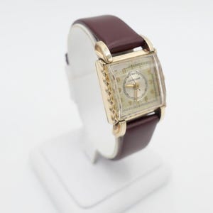 Puede incluir: Un reloj de pulsera Longines de tono dorado con una esfera cuadrada y una correa de cuero marrón. El reloj está en un expositor blanco.