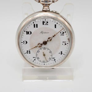 May include: A silver Alpina pocket watch with a white face, black numerals, and a subdial. The watch has a silver case and a gold-coloured crown. The watch is displayed on a clear stand. The number 2690 is printed on a white label.