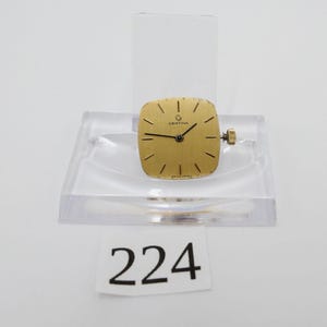 May include: A gold Certina watch face with black hands and markers, presented on a clear acrylic stand. The watch features a square face and a small crown. The number "224" is visible on a white tag below the stand.