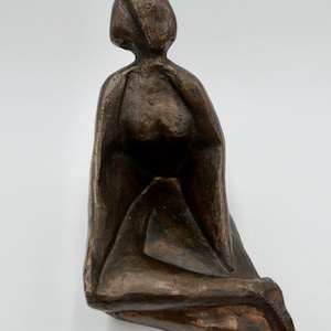 Könnte beinhalten: Eine Bronzeskulptur einer sitzenden Figur in einem vereinfachten, abstrakten Stil. Die Figur ist mit einem drapierten Gewand dargestellt und hat eine kontemplative Pose.
