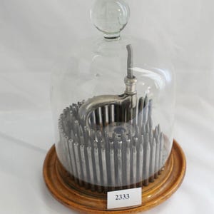 May include: A glass dome with a wooden base and a metal device inside. The device has a handle and a series of metal rods. The base has a label with the number 2333.