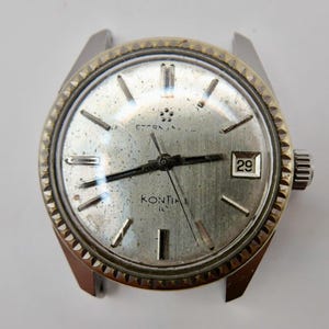 Puede incluir: Un reloj plateado vintage con bisel dorado. La esfera muestra el texto "ETERNA" y "KONTIKI" con manecillas y marcadores negros. La fecha es visible a las 3 en punto. El diámetro del reloj es de aproximadamente 3,5 cm.